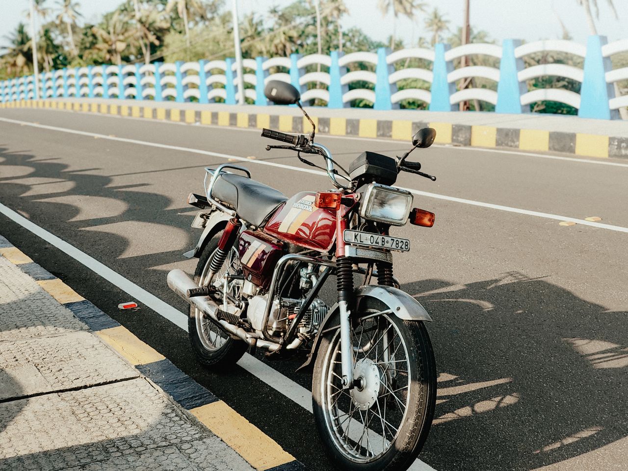A vintage motorcycle parked on a sunlit road in Cherthala, Kerala, with a classic bridge in the background.